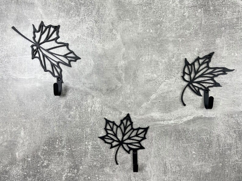May include: Three black metal leaf-shaped hooks on a gray textured wall. Each hook is shaped like a maple leaf with a rounded hook at the bottom.