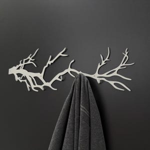 May include: A white metal wall-mounted coat rack shaped like a tree branch. A gray garment hangs from the rack against a dark gray wall. The rack has multiple branching points for hanging items.