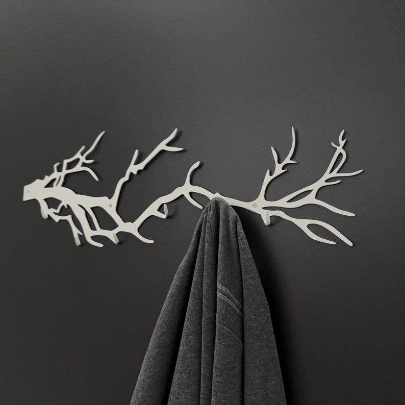 May include: A white metal wall-mounted coat rack shaped like a tree branch. A gray garment hangs from the rack against a dark gray wall. The rack has multiple branching points for hanging items.