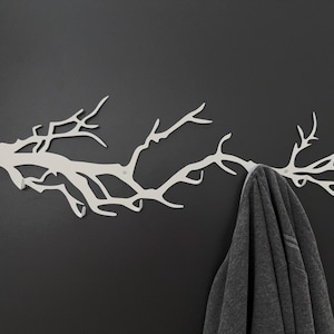 May include: A white metal wall-mounted coat rack shaped like a tree branch. The rack is mounted on a dark gray wall, and a gray garment hangs from one of the branches. The design is modern and minimalist.