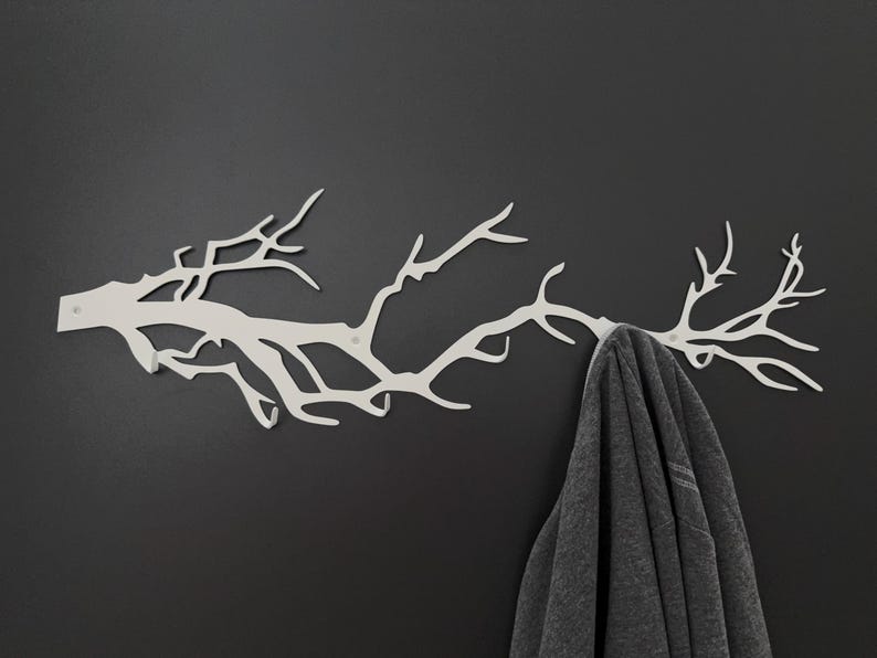 May include: A white metal wall-mounted coat rack shaped like a tree branch. The rack is mounted on a dark gray wall, and a gray garment hangs from one of the branches. The design is modern and minimalist.