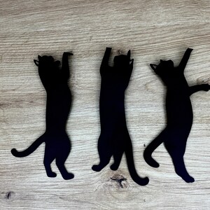 Pu&ograve; includere: Tre sagome di gatti neri ritagliate su carta e disposte su uno sfondo di legno. I gatti sono in varie pose, con un gatto in piedi sulle zampe posteriori, un gatto seduto e un gatto sdraiato.