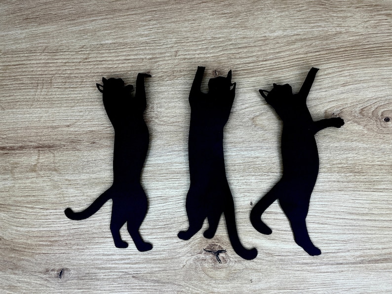 Pu&ograve; includere: Tre sagome di gatti neri ritagliate su carta e disposte su uno sfondo di legno. I gatti sono in varie pose, con un gatto in piedi sulle zampe posteriori, un gatto seduto e un gatto sdraiato.