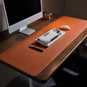 Pode incluir: Um tapete de mesa de couro castanho com uma borda costurada, um teclado branco com teclas cinzentas e pretas, um rato de computador e um porta-canetas numa secretária de madeira. Um monitor de computador e uma chávena pequena também são visíveis.