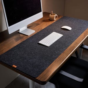 Puede incluir: Una alfombrilla de escritorio de fieltro gris oscuro sobre un escritorio de madera. Un teclado y un ratón blancos están sobre la alfombrilla. Un monitor de ordenador y una pequeña taza de madera con una cuchara también están sobre el escritorio. El escritorio tiene un acabado de madera oscura.