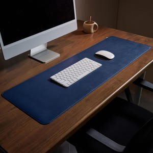 May include: A navy blue desk mat with a white keyboard and mouse on a wooden desk. The mat is rectangular and covers a large area. A computer monitor and a small wooden cup are also present.