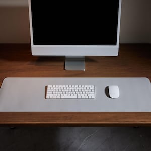 Könnte beinhalten: Eine hellgraue rechteckige Schreibtischunterlage mit einer weißen Tastatur und Maus. Ein silberner Computermonitor befindet sich im Hintergrund. Die Schreibtischunterlage liegt auf einem dunkelbraunen Holzschreibtisch.