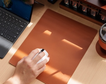 May include: A brown leather mousepad with a white wireless mouse on top. The mousepad is on a light wood surface.