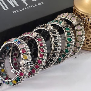 Op de afbeelding: Een verzameling zilverkleurige armbanden met kleurrijke edelsteenaccenten. De armbanden hebben verschillende vormen en kleuren, waaronder rood, groen, roze en blauw. De sieraden worden gepresenteerd in de buurt van een zwarte doos met de tekst "THE LIFESTYLE HUB".