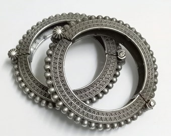 May include: Two silver bangles with intricate designs and beaded edges. The bangles are open and hinged, allowing them to be easily put on and taken off.