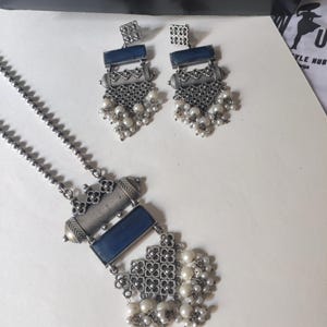 May include: A silver-toned jewelry set featuring a necklace and matching earrings. The necklace has a rectangular blue stone pendant with floral accents and a cluster of white pearls. The earrings have a similar design, with square tops and dangling pearls.