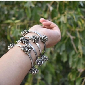 Puede incluir: Un conjunto de cuatro pulseras de plata con racimos decorativos de pequeñas bolas de plata. Las pulseras están apiladas en la muñeca de una persona.