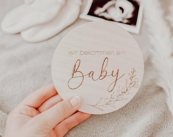 May include: A wooden round sign with the text "wir bekommen ein Baby" in cursive script, surrounded by a floral design. The sign is held in a hand against a light brown background.