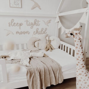 May include: A white wooden toddler bed with a white and brown giraffe stuffed animal, a white goose stuffed animal, and a brown knitted blanket. The bed has a white canopy and a white flag garland. The wall behind the bed has wooden cutouts that say "sleep tight moon child".