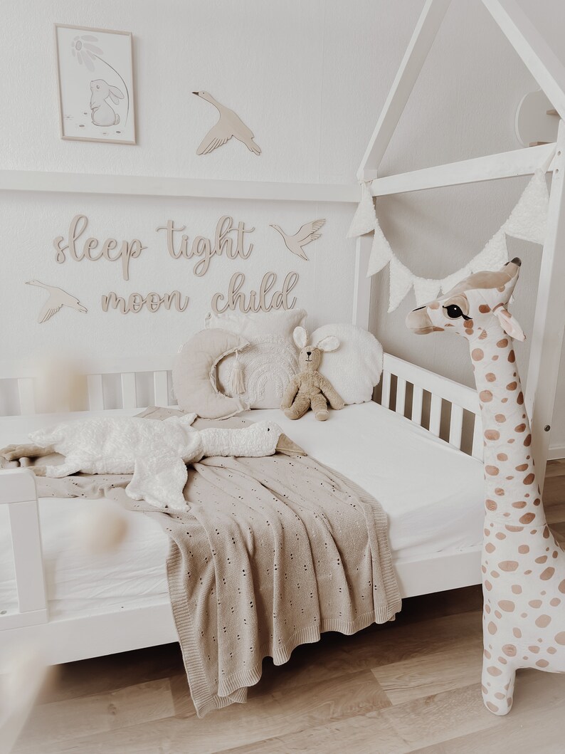 May include: A white wooden toddler bed with a white and brown giraffe stuffed animal, a white goose stuffed animal, and a brown knitted blanket. The bed has a white canopy and a white flag garland. The wall behind the bed has wooden cutouts that say "sleep tight moon child".