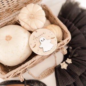 Könnte beinhalten: Ein rundes Holzornament mit einem weißen Geister-Ausschnitt, dem Namen Aurora und dem Text Happy Halloween, verziert mit kleinen Sternen und Fledermäusen. Das Ornament liegt in einem geflochtenen Weidenkorb neben kleinen weißen Kürbissen.