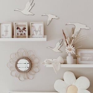 May include: A nursery room with a white changing table, a white shelf with a wooden swan, a white flower pillow, and a wooden sign that says "You are magic".