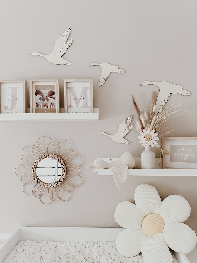 May include: A nursery room with a white changing table, a white shelf with a wooden swan, a white flower pillow, and a wooden sign that says "You are magic".
