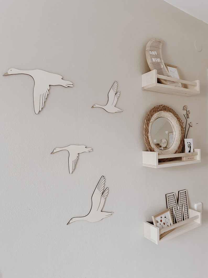 May include: A white wall with four wooden bird cutouts in the shape of geese. There are three wooden shelves with a moon-shaped sign that says "Good Night" and a round mirror with a woven frame.