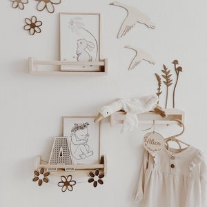 May include: A nursery room decor set with a wooden shelf, a wooden hanger, and a wooden name sign that says "Alica". The shelf has two prints, one of a bunny and one of a baby. The hanger has a stuffed goose toy hanging from it. The room is decorated with wooden flowers and geese.