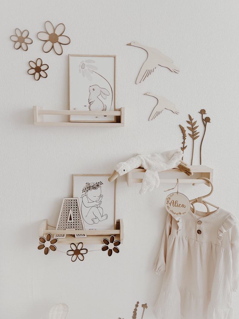 May include: A nursery room decor set with a wooden shelf, a wooden hanger, and a wooden name sign that says "Alica". The shelf has two prints, one of a bunny and one of a baby. The hanger has a stuffed goose toy hanging from it. The room is decorated with wooden flowers and geese.