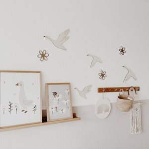 May include: A white wall with wooden wall decor featuring geese and flowers. Two framed prints, one with a goose and the other with a girl in a field of flowers, are on a wooden shelf. A wooden rainbow wall hanging and a woven basket are on a wooden rack.