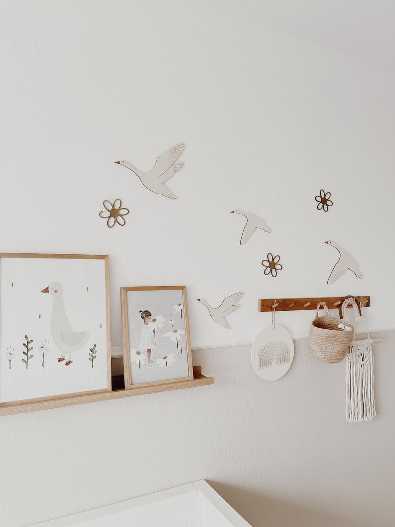May include: A white wall with wooden wall decor featuring geese and flowers. Two framed prints, one with a goose and the other with a girl in a field of flowers, are on a wooden shelf. A wooden rainbow wall hanging and a woven basket are on a wooden rack.