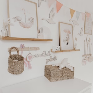 May include: A white dresser with a wicker basket on top. The basket has a stuffed animal on top of it. The wall behind the dresser is decorated with wooden animal cutouts, a wooden sign that says "unendlich geliebt", and a string of pastel colored flags.