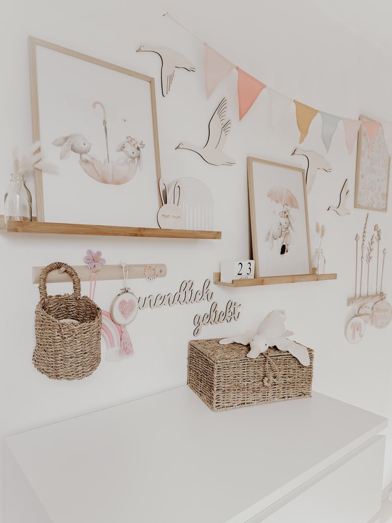 May include: A white dresser with a wicker basket on top. The basket has a stuffed animal on top of it. The wall behind the dresser is decorated with wooden animal cutouts, a wooden sign that says "unendlich geliebt", and a string of pastel colored flags.