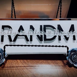 Pode incluir: Uma placa branca com a palavra "GRANDMA" em letras pretas. Os nomes "Kathy", "Helene" e "Magan" estão escritos em itálico em uma fita branca acima da palavra "GRANDMA". Duas flores de papel azuis estão de cada lado da placa.