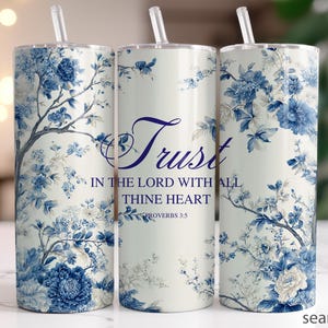 May include: Three cylindrical tumblers with clear straws, featuring a white background and a blue floral pattern. The center tumbler displays the text Trust IN THE LORD WITH ALL THINE HEART PROVERBS 3:5 in a dark blue script and serif font.
