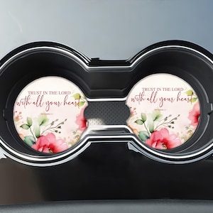 May include: Two round car coasters with floral designs and the text "Trust in the Lord with all your heart." The coasters feature pink flowers and green leaves on a light background, set in a car's cup holder.
