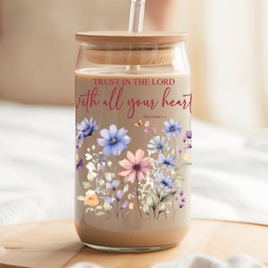 Puede incluir: Un vaso de vidrio transparente con tapa de madera y pajita. El vaso está lleno de un líquido marrón claro y decorado con un diseño floral y el texto "Trust in the Lord with all your heart Proverbs 3:5".