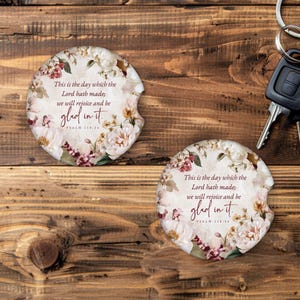 May include: Two floral car coasters with the text "This is the day which the Lord hath made; we will rejoice and be glad in it." The coasters feature a white background with pink and cream-colored flowers. Keys are in the background.