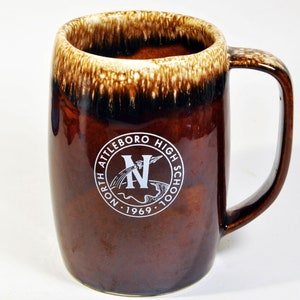 May include: Brown and white glazed ceramic mug with a white logo that reads "North Attleboro High School" and a stylized "N" with a rocket and a river. The logo also includes the year "1969".