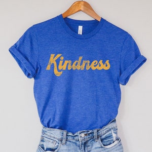 Op de afbeelding: Een blauw T-shirt met het woord "Kindness" in geel op de voorkant. Het shirt hangt aan een houten hanger en wordt gecombineerd met een spijkerbroek.