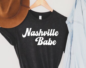 Puede incluir: Una camiseta gris oscuro con el texto "Nashville Babe" en letras blancas de estilo retro. La camiseta está anudada en la parte inferior y colgada de una percha de madera. También hay un sombrero marrón y una chaqueta vaquera azul claro en la imagen.