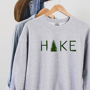 Op de afbeelding: Grijs sweatshirt met het woord "HAKE" bedrukt in groene letters met een groene dennenboom die de "A" vervangt.