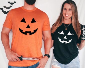 May include: A couple wearing matching Halloween themed t-shirts. The man is wearing an orange t-shirt with a black jack-o'-lantern face. The woman is wearing a black t-shirt with a white jack-o'-lantern face.