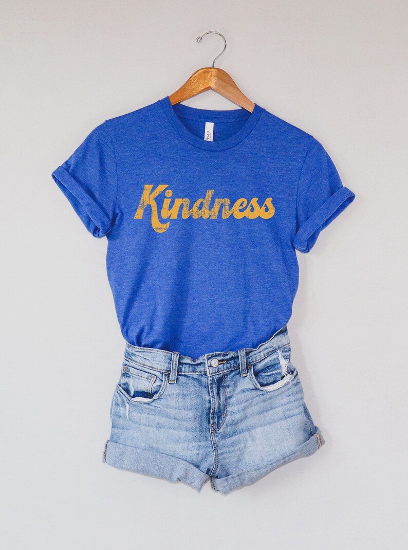 Op de afbeelding: Een blauw T-shirt met het woord "Kindness" in geel op de voorkant. Het shirt hangt aan een houten hanger en wordt gecombineerd met een spijkerbroek.
