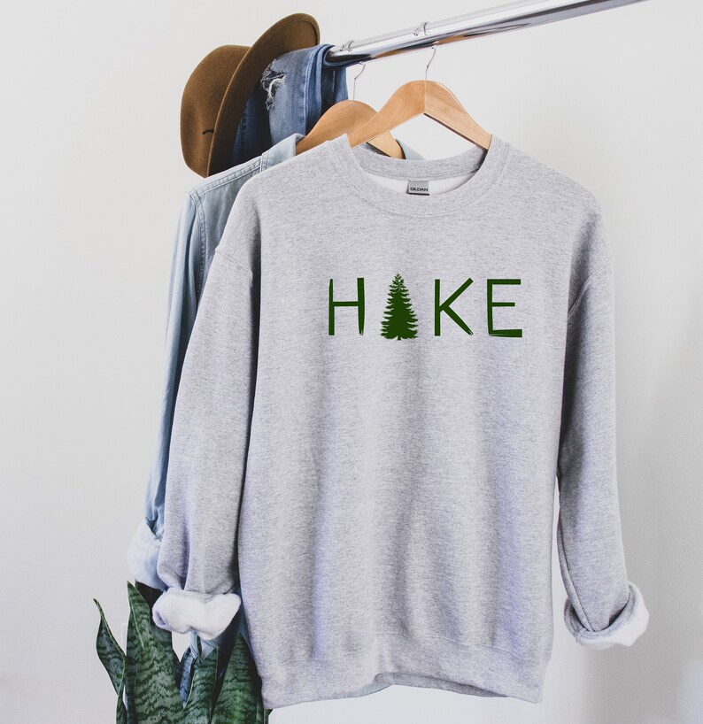 Op de afbeelding: Grijs sweatshirt met het woord "HAKE" bedrukt in groene letters met een groene dennenboom die de "A" vervangt.
