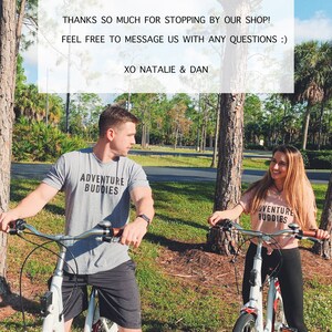 Op de afbeelding: Twee mensen fietsen op een pad in een park. Ze dragen allebei T-shirts met de tekst "Adventure Buddies". De tekst "THANKS SO MUCH FOR STOPPING BY OUR SHOP! FEEL FREE TO MESSAGE US WITH ANY QUESTIONS :) XO NATALIE & DAN" wordt weergegeven op een witte achtergrond.