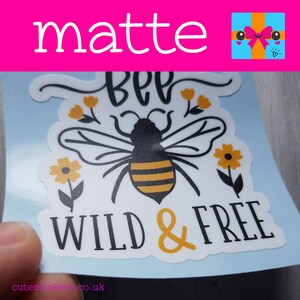 May include: A matte sticker featuring a bee illustration with the words "BEE WILD & FREE" in black and yellow. The bee has black and yellow stripes, with yellow flowers and leaves. The sticker is on a light blue background.
