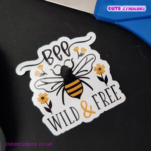 May include: A white sticker with a black and yellow bee illustration, the words "BEE WILD & FREE", and small yellow flower accents. The sticker is on a black surface with blue scissors in the background. The text "Cute Stickers" is visible.