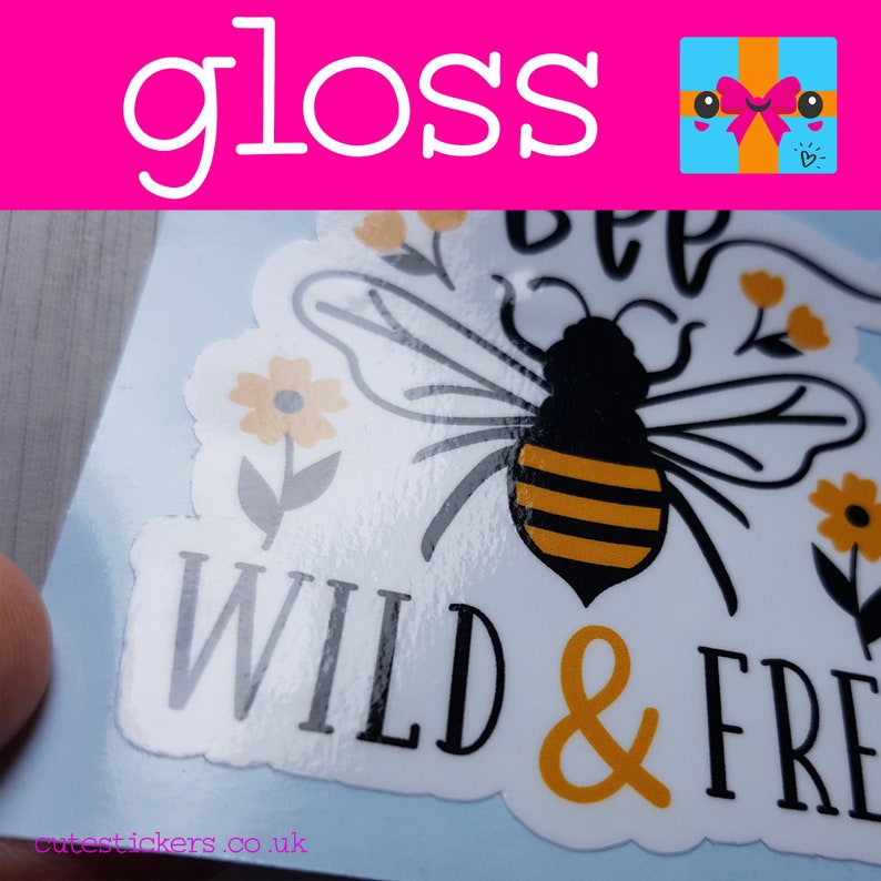 May include: A glossy sticker with a bee illustration and the words "BEE WILD & FREE" in black and orange. The sticker also features yellow flowers and the text "gloss" in white on a pink background.
