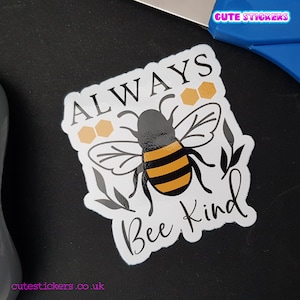 May include: A white sticker with a black and yellow bee illustration, the words "ALWAYS Bee Kind" in a handwritten font, and honeycomb graphics. The sticker has a glossy finish and is on a black surface. The text "Cute Stickers" is in the top right corner.
