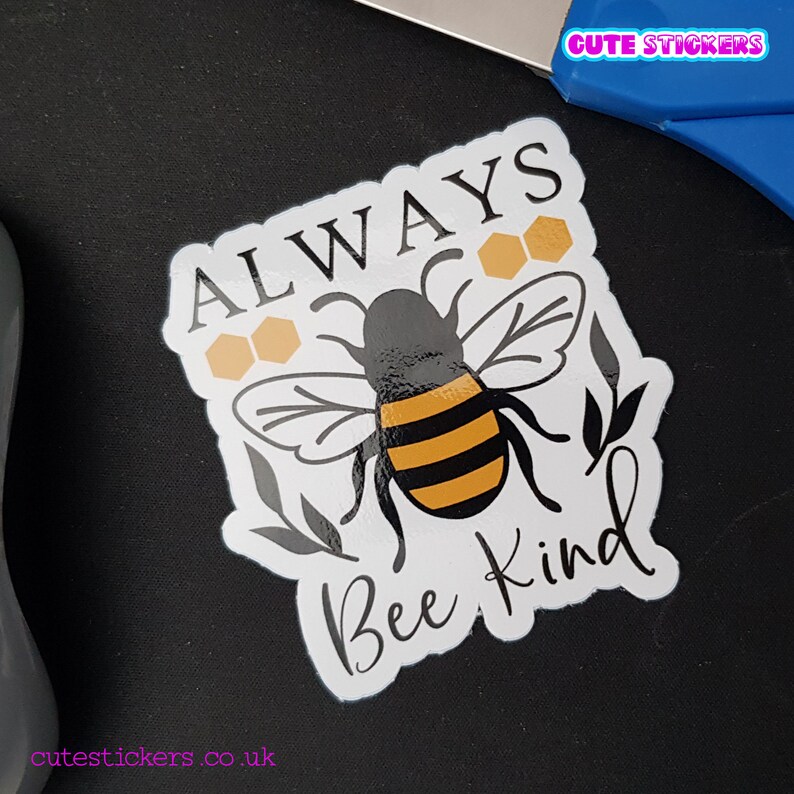 May include: A white sticker with a black and yellow bee illustration, the words "ALWAYS Bee Kind" in a handwritten font, and honeycomb graphics. The sticker has a glossy finish and is on a black surface. The text "Cute Stickers" is in the top right corner.