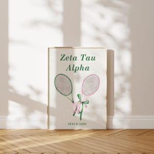May include: A framed print featuring two watercolor tennis rackets, one green and one pink, tied with a green and pink bow. The text "Zeta Tau Alpha" and "SINCE 1898" are displayed in green. The background is a neutral off-white.