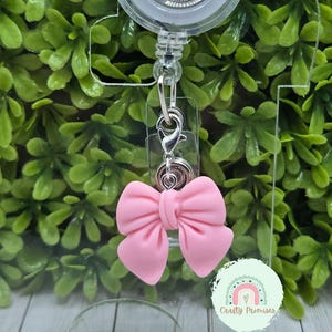 Può includere: Un porta badge retrattile con una clip argentata e un ciondolo a forma di fiocco rosa. Il ciondolo è attaccato a una catena argentata. Il porta badge è in plastica trasparente e ha una clip girevole argentata. Lo sfondo è una pianta verde frondosa.