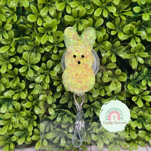 May include: A yellow and green glitter peep bunny charm attached to a clear retractable badge holder. The holder has a silver clip and a silver swivel hook. The charm is attached to the holder with a silver ring. The background is a green leafy plant.